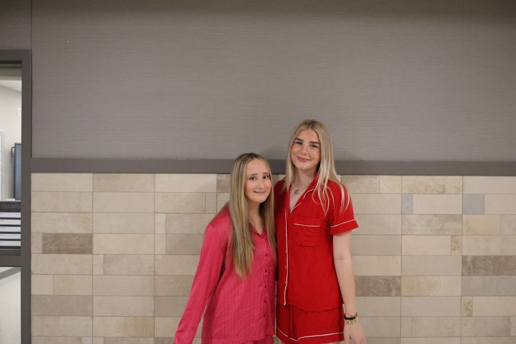 Two women in pajamas, one in pink and the other in red, stand together before a tiled wall.