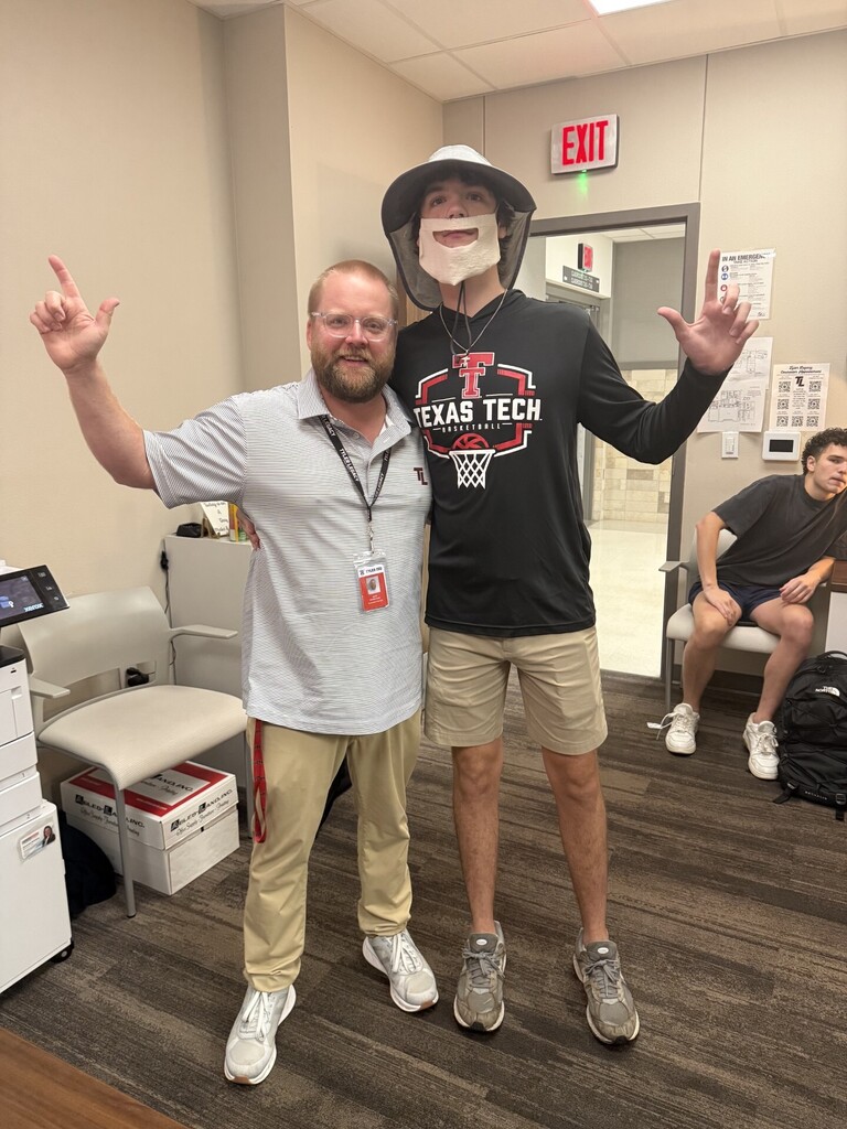 Two men pose in an office with one wearing a mask. A desk and chairs are behind them.