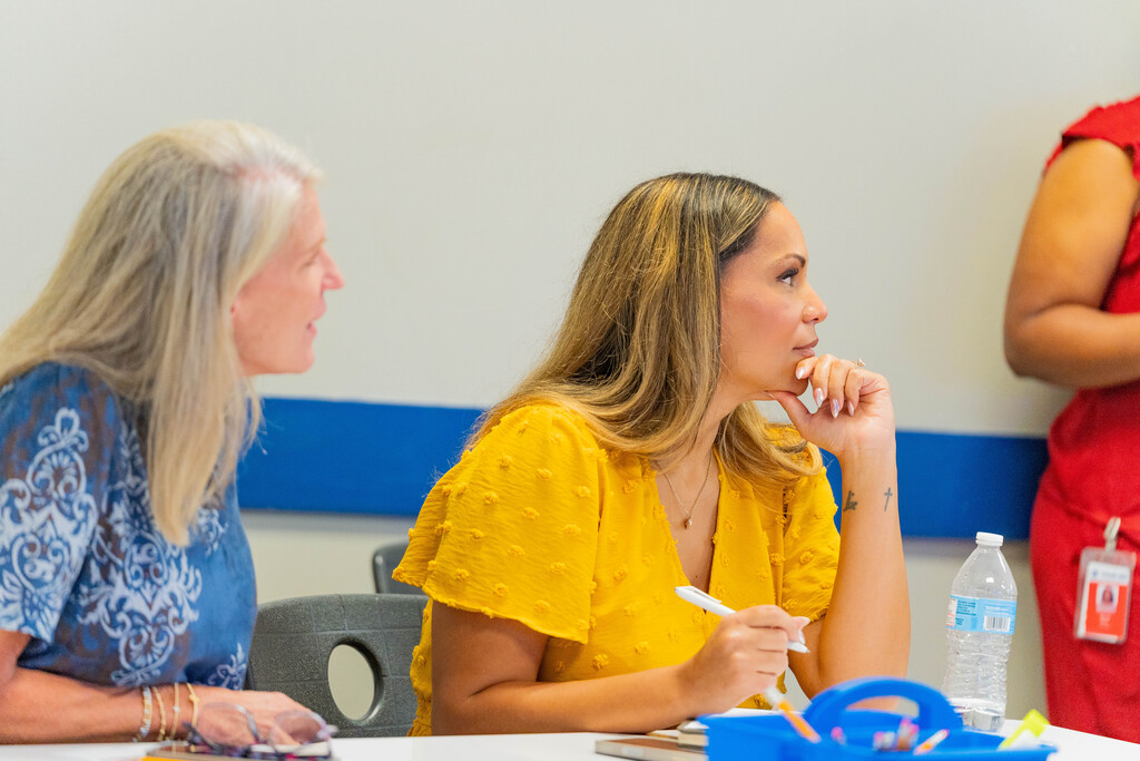 Two women converse at a table, one in a blue dress and the other in yellow, with a third standing nearby.
