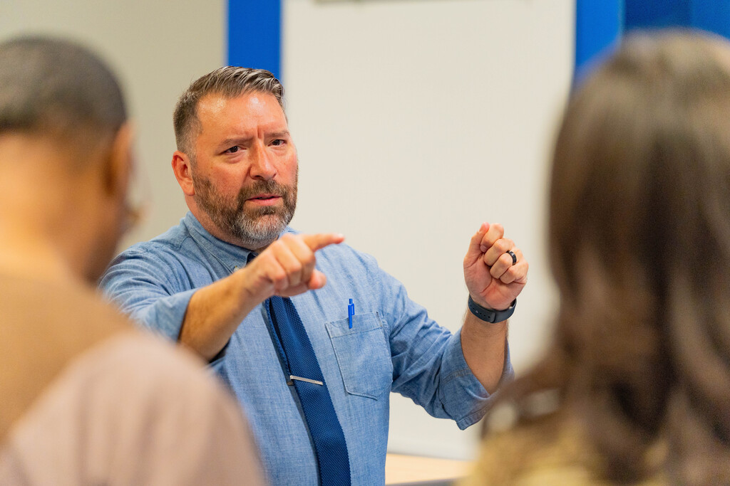 A man speaks to two others in a room. He points with one hand and gestures with the other.
