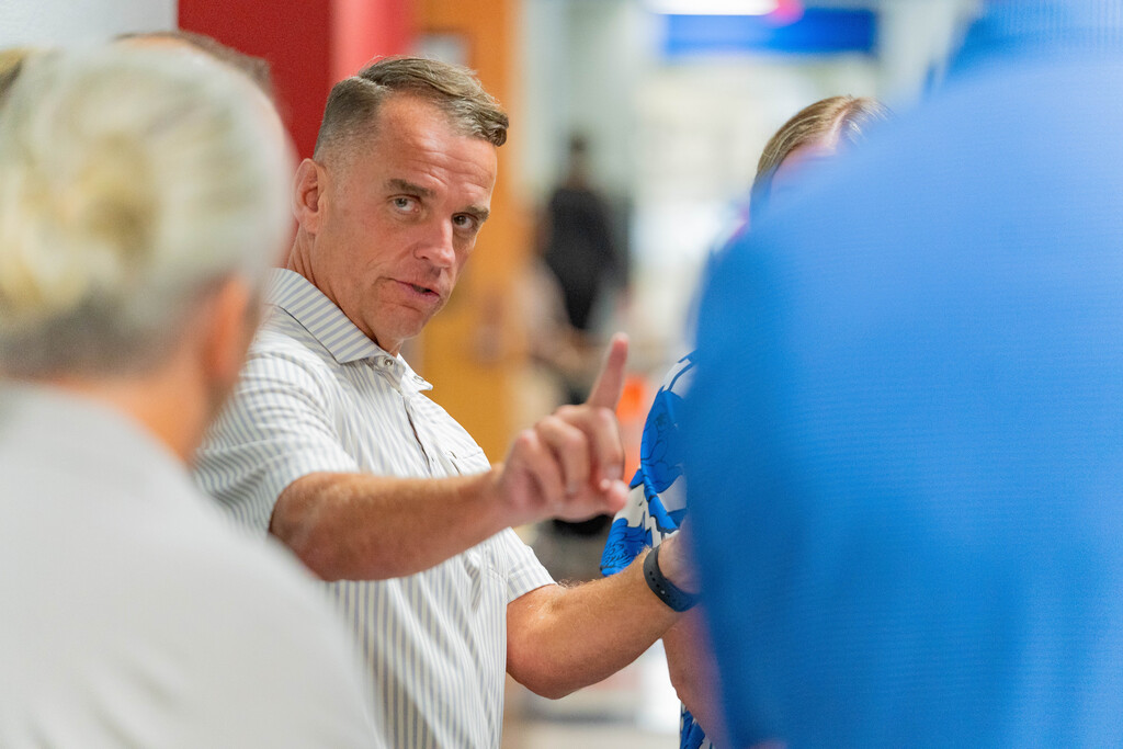 A man speaks to others in a hallway, gesturing with his hand, surrounded by others in a busy area.