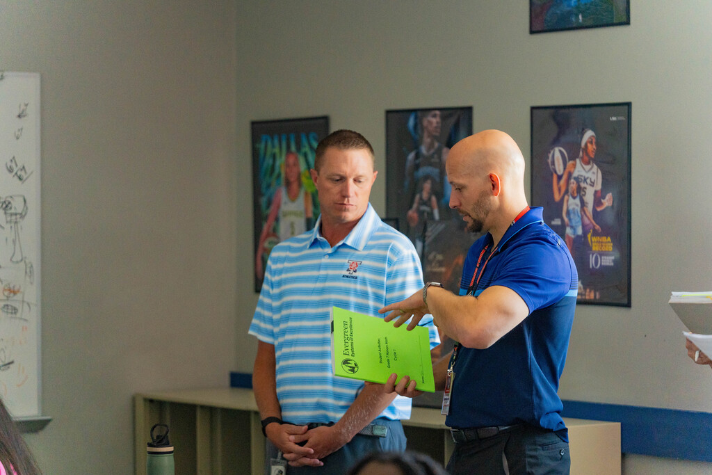 Two men, one holding a green paper, stand in a room with white walls and framed pictures.