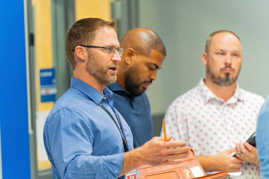 Three men in an indoor setting, one in a blue shirt speaks, another in a patterned shirt holds a phone.