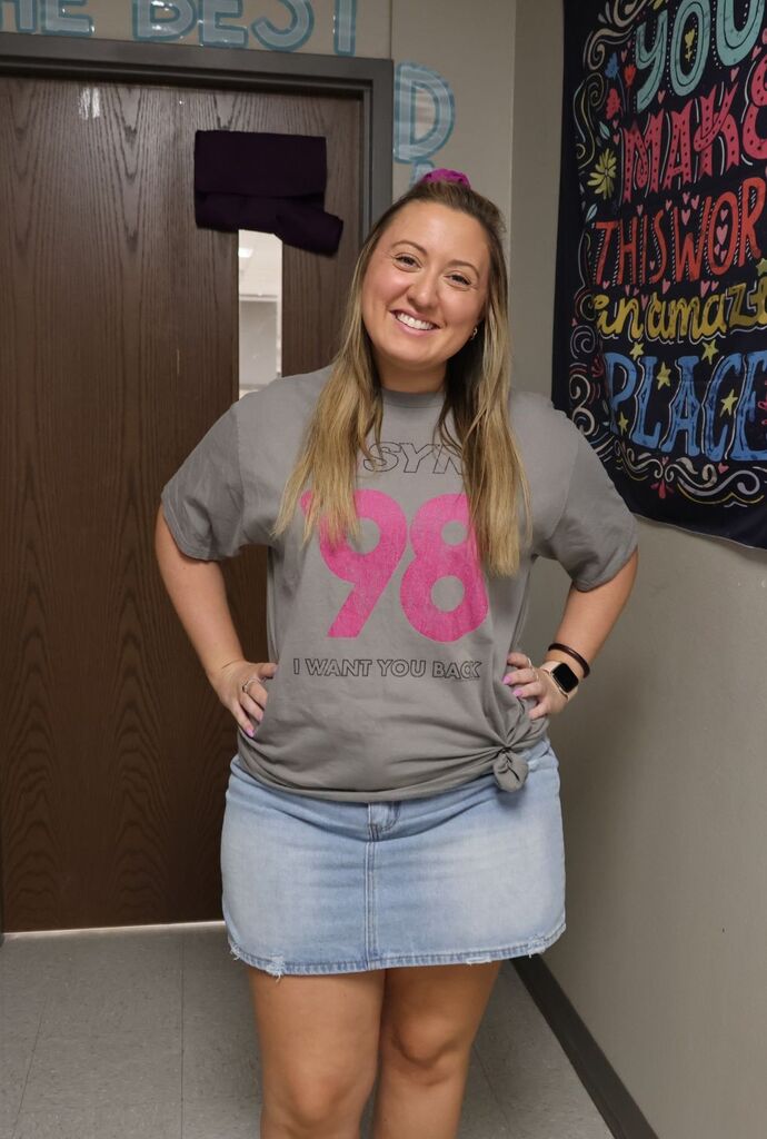 Woman in a gray tee shirt with pink 98, a denim skirt, and a bracelet stands smiling in a hallway with a brown door and a motivational poster.