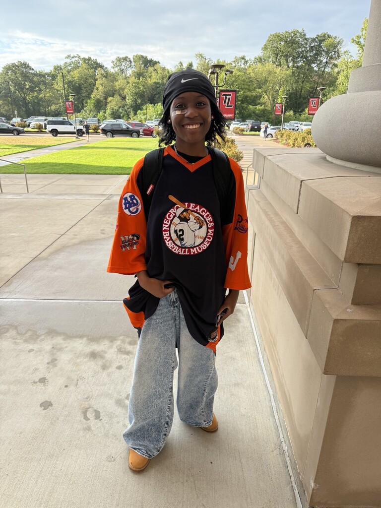 Person in orange and black jersey with 'New York Yankees' logo stands on campus sidewalk. Background includes trees and parked cars.