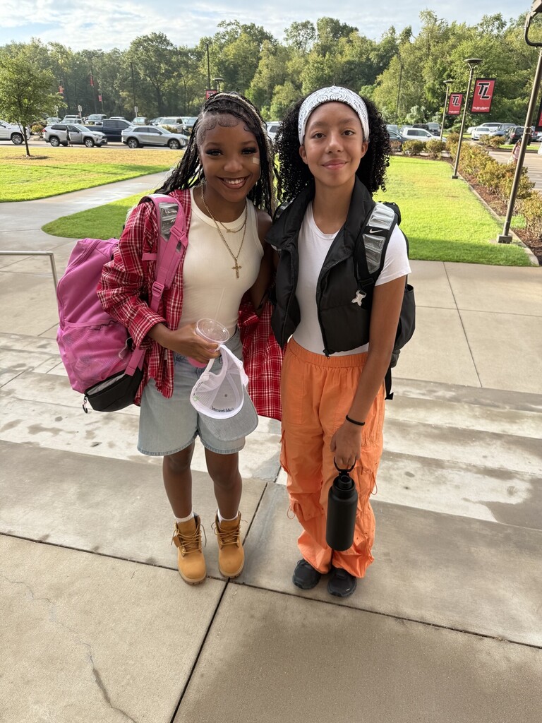 Two children on a sidewalk with backpacks, one in orange pants and the other in shorts, posing for a photo.