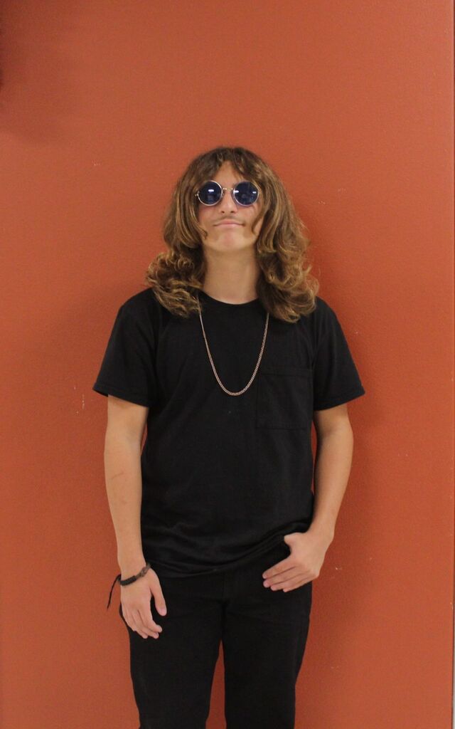 Person with long hair and sunglasses stands against a red wall, wearing black clothes and a necklace.