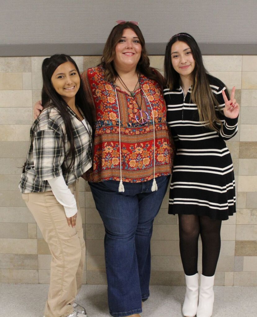 Three women in casual clothes stand close to each other. The one in the middle has a necklace.