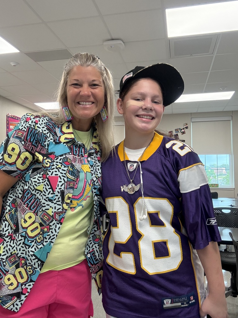 A woman and a boy are standing close together in a room. The woman wears a colorful shirt with a necklace. The boy wears a purple football jersey.