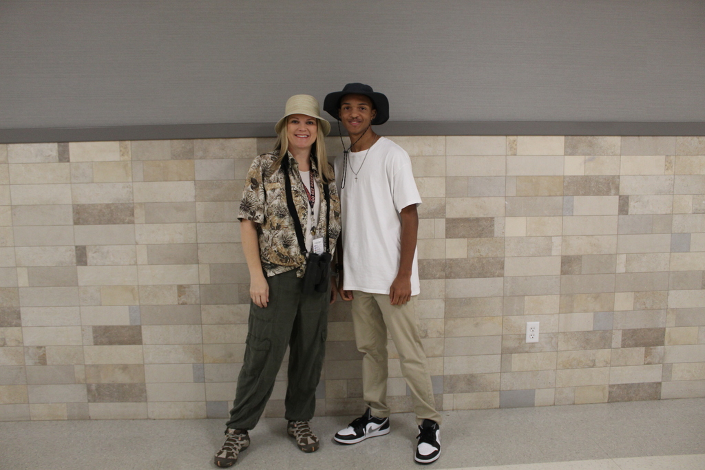 Two people stand next to each other in front of a tiled wall. One wears a hat and a camouflage top.