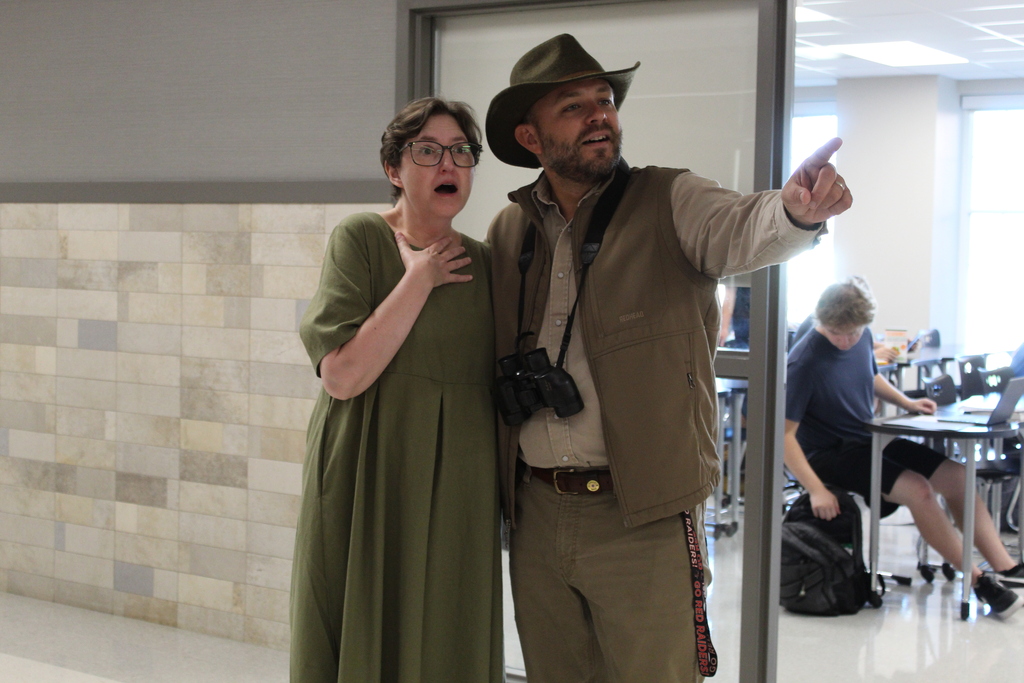 A man in a safari hat and a woman in a green dress point at each other. Behind them is a patterned wall and a seated person.