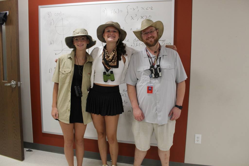Three people wearing safari-style hats pose for a photo in front of a whiteboard.