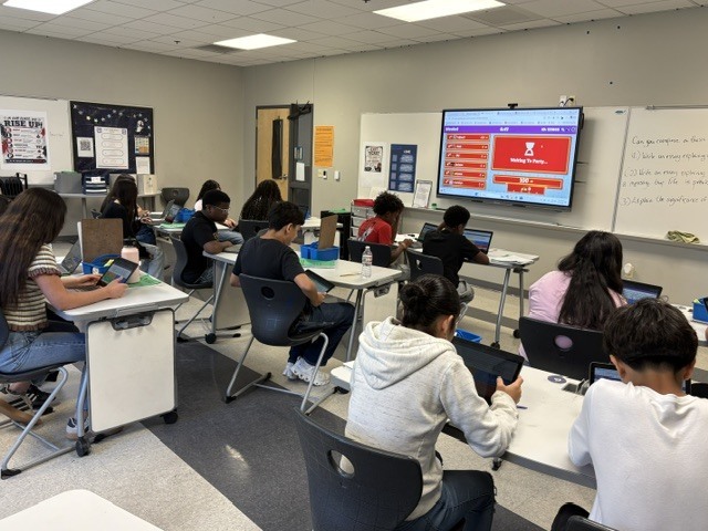 Students using laptops in a classroom with a large monitor on the wall and white boards.