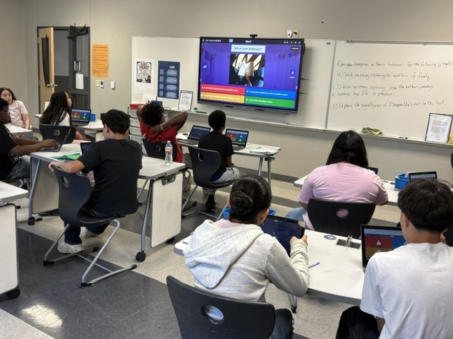 A classroom with students sitting at desks, each using laptops. A screen is projected on a whiteboard.