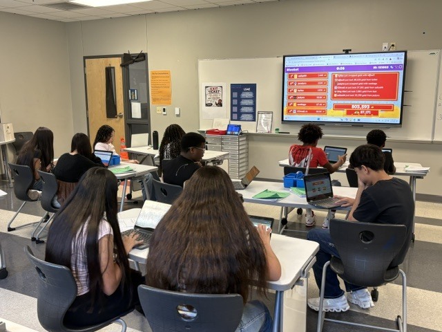 A classroom with students seated at tables, using laptops and looking at a screen displaying information.