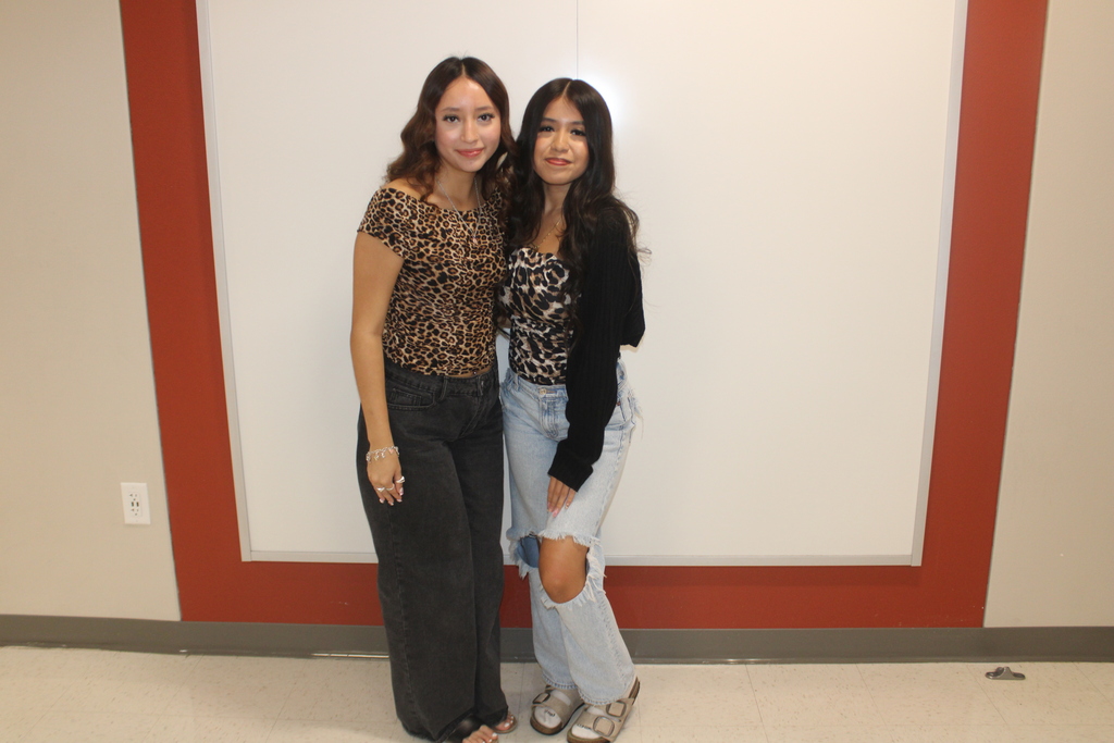 Two women pose for a photo. One wears a leopard-print top and jeans. The other wears a black sweater and ripped jeans.