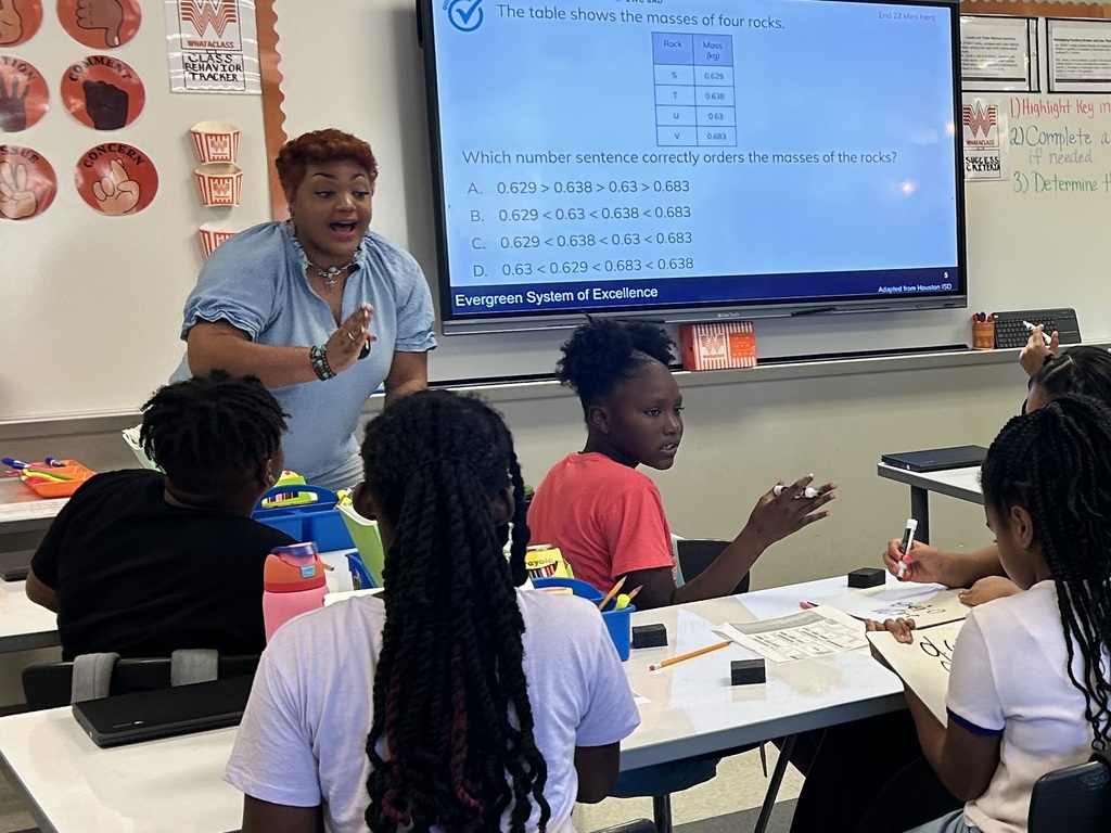 A teacher in a blue shirt stands before a classroom, addressing students. Students sit at tables, engaging with materials and one holds a pen.