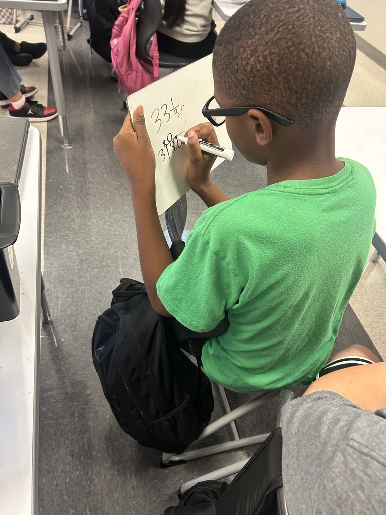 A boy wearing a green shirt and glasses writes on a white paper inside a classroom.