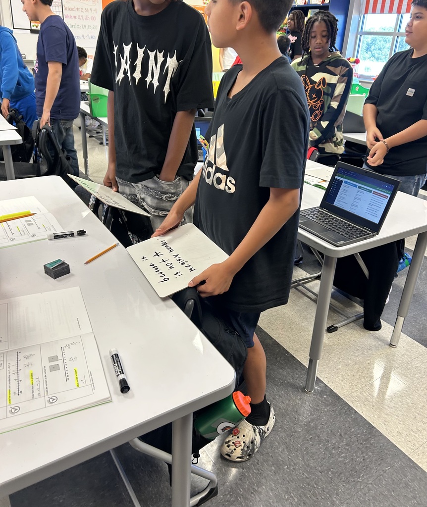 A group of students in a classroom. One student holds a paper, another holds a laptop, and others are standing.