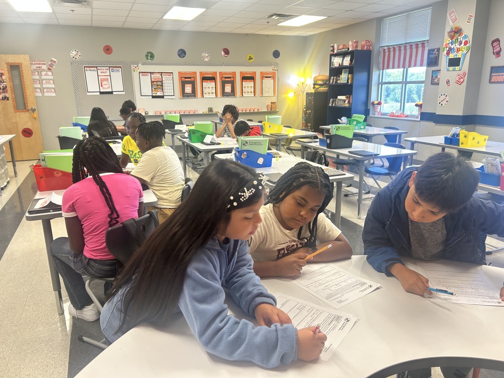 Several students work at desks in a classroom with a white ceiling and bulletin boards.