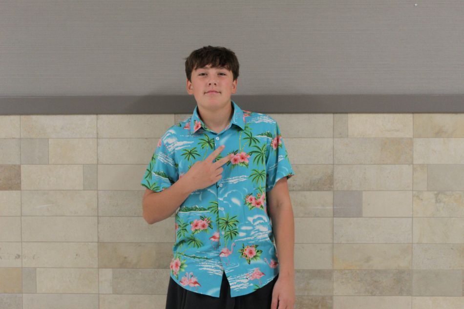 A boy in a blue floral shirt gestures with his left hand in front of a tiled wall.