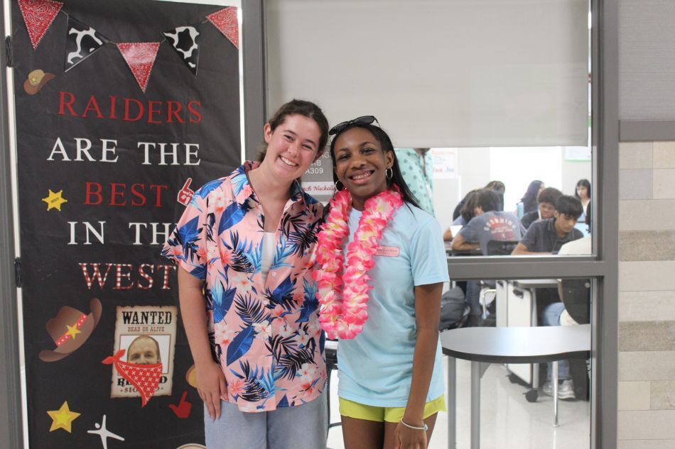 Two women standing by a wall with a sign. One wears a floral shirt and lei; the other wears a turquoise shirt.