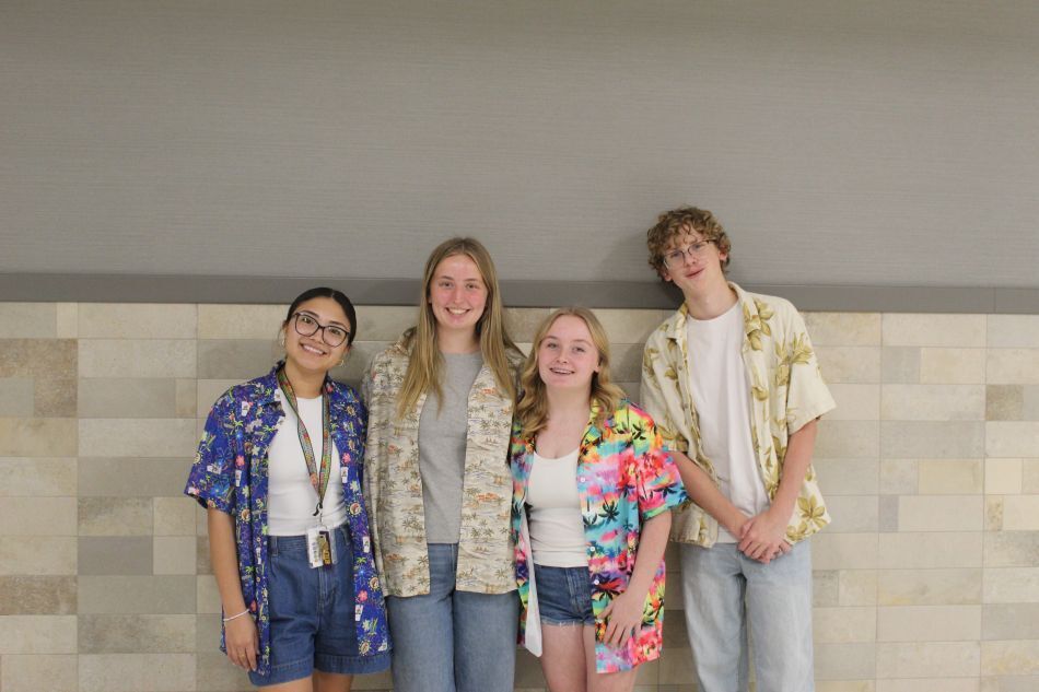Four people stand together, smiling, with tiled wall in the background. They wear casual summer clothing.