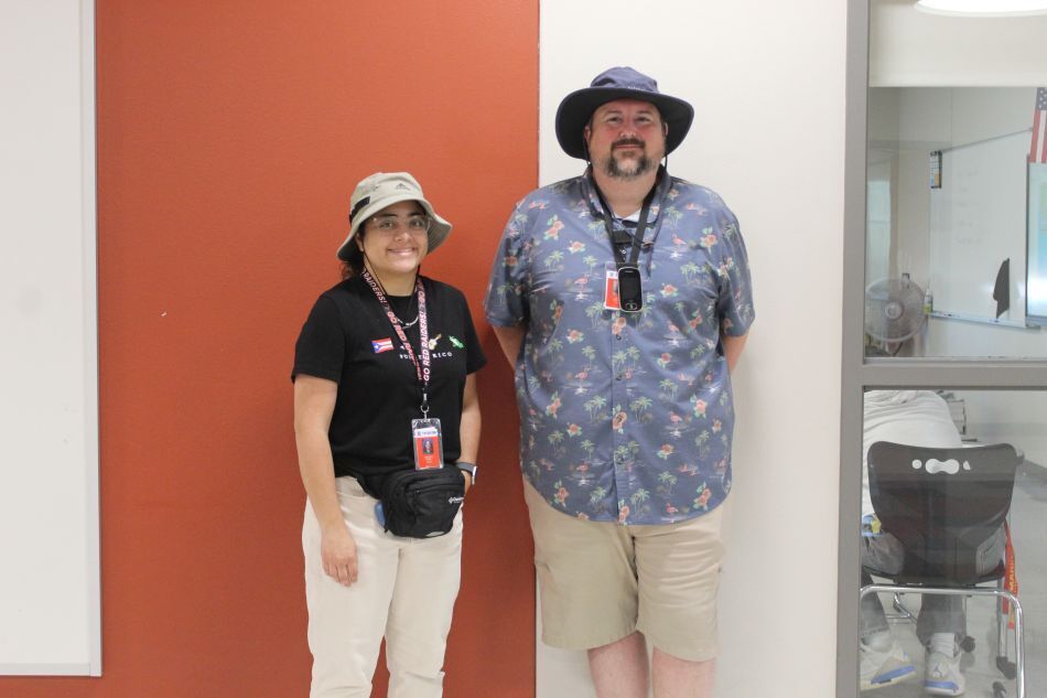 A man and a woman pose together. The man wears a blue shirt and khaki shorts. The woman wears a black shirt, hat, and white pants.
