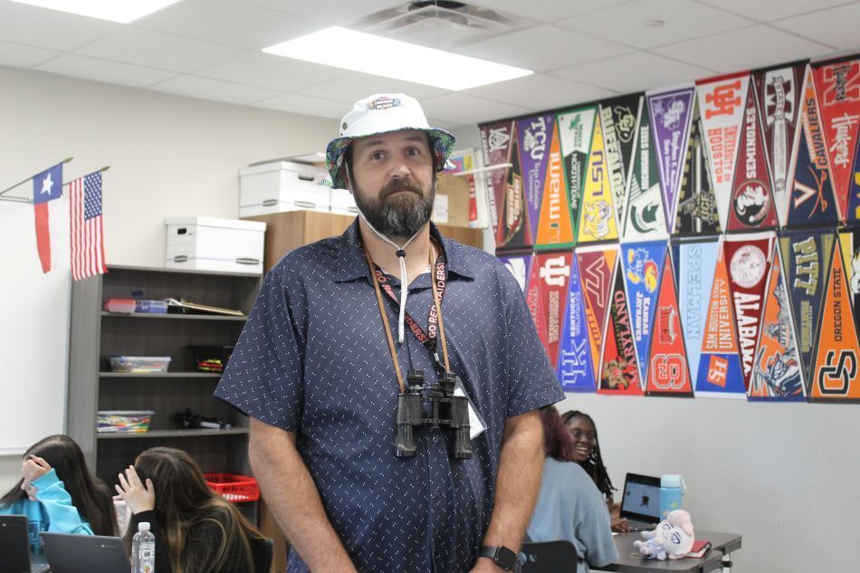 A man with a white hat stands in a room with bookshelves and students, wearing binoculars.