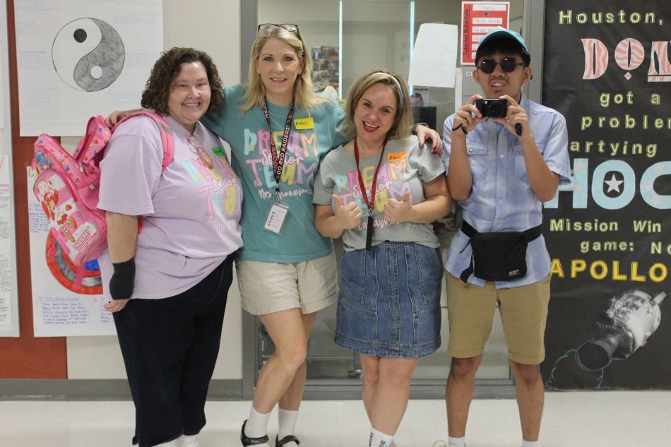 Four people stand in a hallway, smiling. One wears sunglasses and a cap. Behind them is a banner with text.