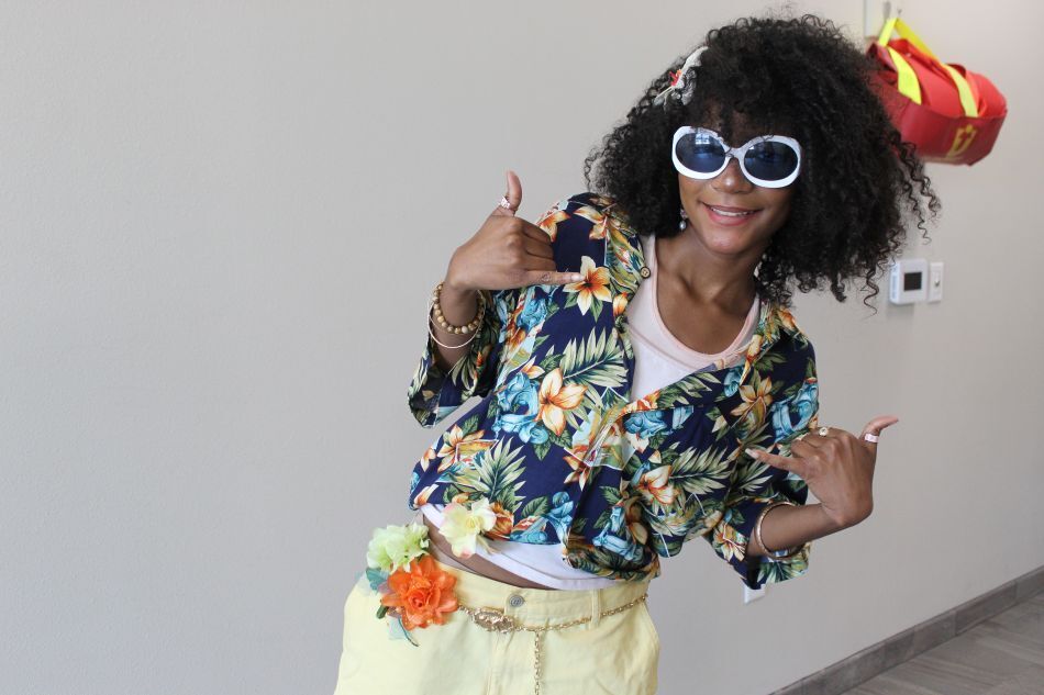 Woman in floral jacket and sunglasses posing with hands up, yellow pants, and background includes white wall and red bag.