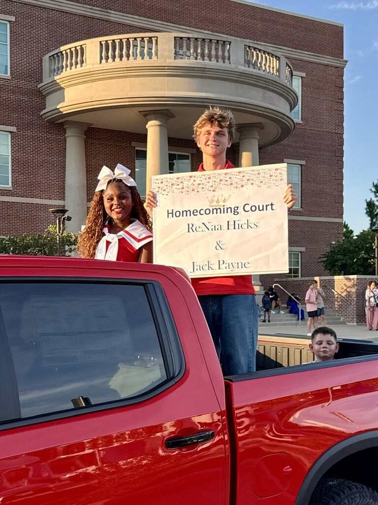 Two people in red uniforms hold a sign for Homecoming Court. A child sits in the truck bed. A building with columns stands in the background.