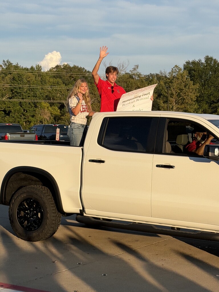 Two people standing in a white pickup truck, one holding a paper, waving at the camera. Trees in the background.