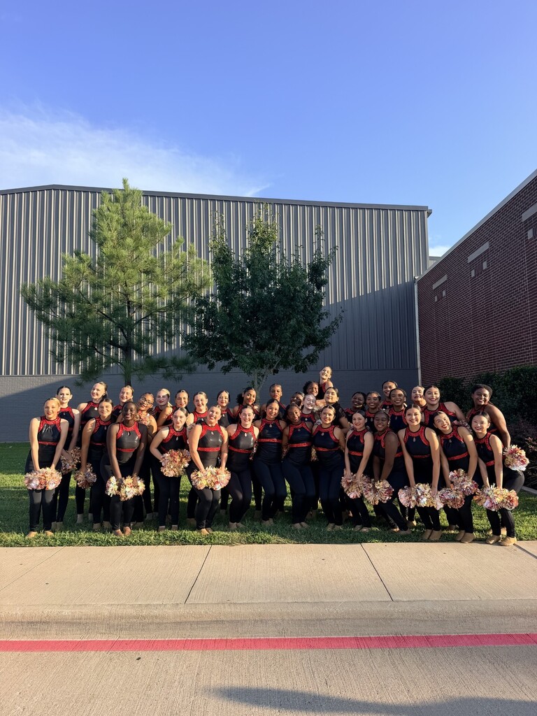 Large group of cheerleaders in red and black uniforms holding pom-poms, standing on grass in front of buildings.