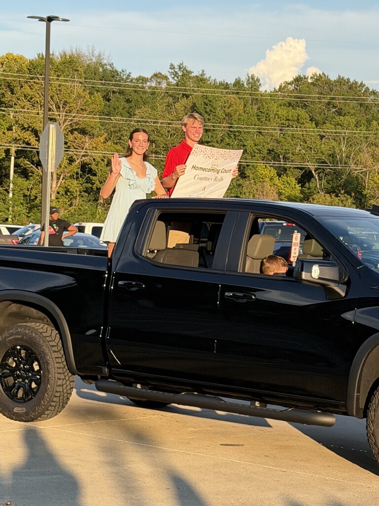 Two people in a pickup truck. One holding a sign. Another waving. Trees and electric poles in the background.