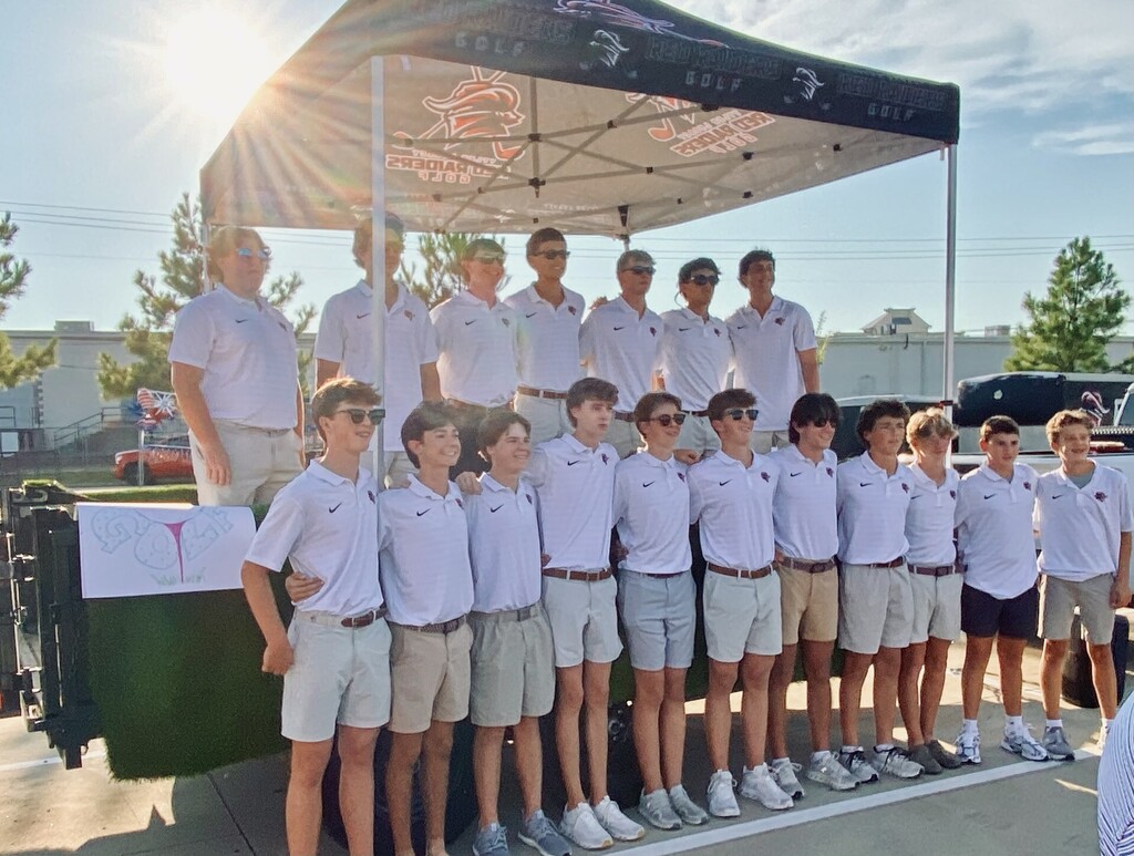 A group of young men wearing white shirts and shorts are standing under a tent. Trees and buildings are visible in the background.