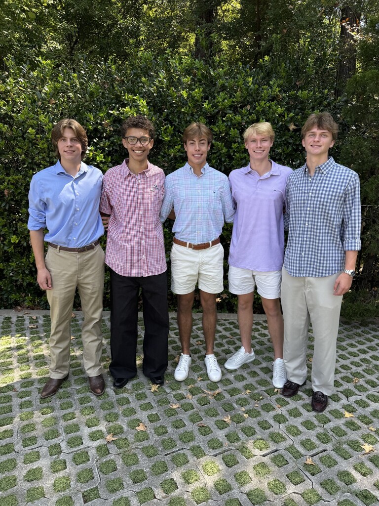 Five males dressed in various colors and styles of shirts and pants standing in front of greenery.
