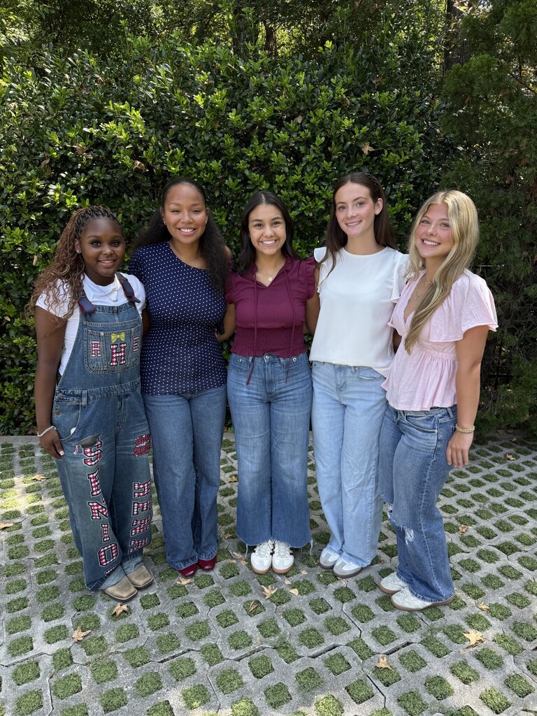Five women stand together on a tiled path, wearing various colored tops and jeans. They pose with greenery in the background.