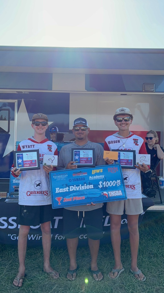 Four individuals stand on grass, holding certificates and a large blue sign. One wears a white hat, one a red hat, and two wear sunglasses.