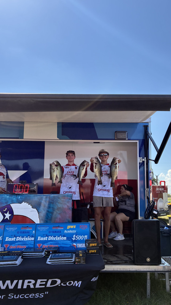 Two individuals holding fish at a competition, one with a backpack, on a stage with a blue backdrop.