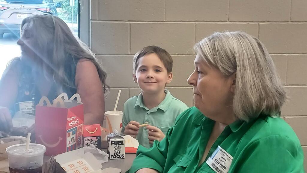 Woman and boy seated at a table, smiling. In front of them, drinks, straws, and food. Blurry background of another woman.