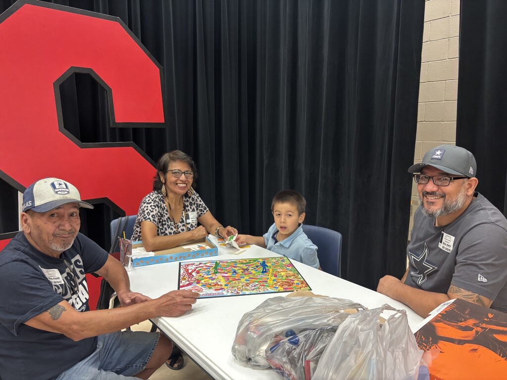 Three adults and a child sit at a table. A large red number eight is behind them.