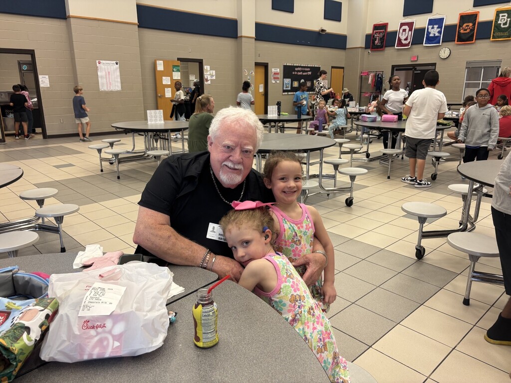 A man in a black shirt sits at a table with two young girls in dresses. They are in a cafeteria with tables and chairs.