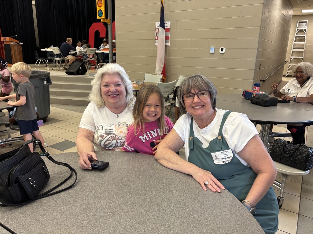 Three women and a child are seated at a table, smiling. The adult women wear glasses and aprons. A black bag and a cellphone are on the table.