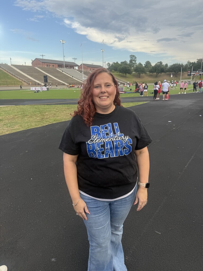 A woman wearing a black t-shirt with the text "BELL PRIMARY BEARS" stands on a track at a stadium.