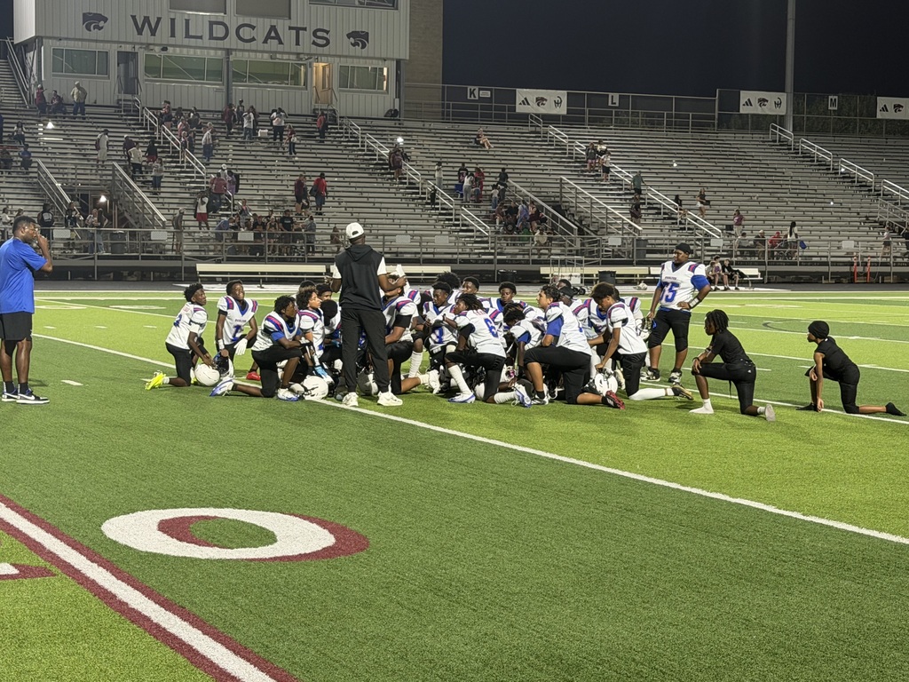 Football team huddled on a field with a "Wildcats" sign at the top of the stands.