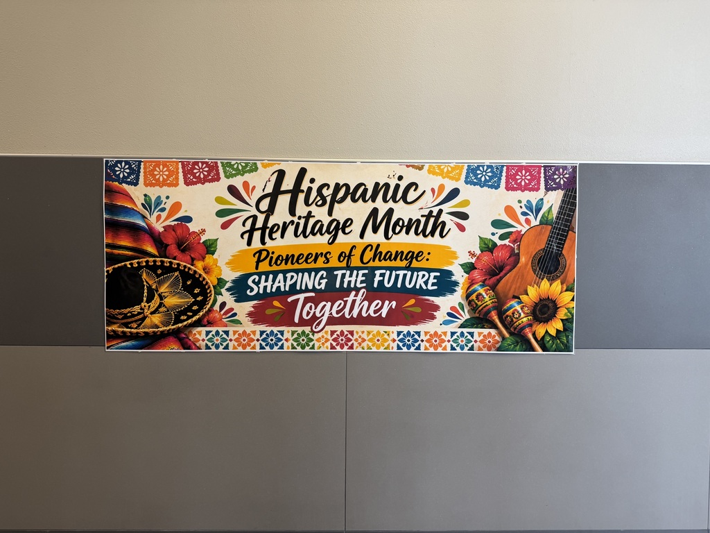 A sign with Hispanic Heritage Month theme mounted on a gray wall. The sign features a sombrero, guitar, flowers, and bold colorful text.