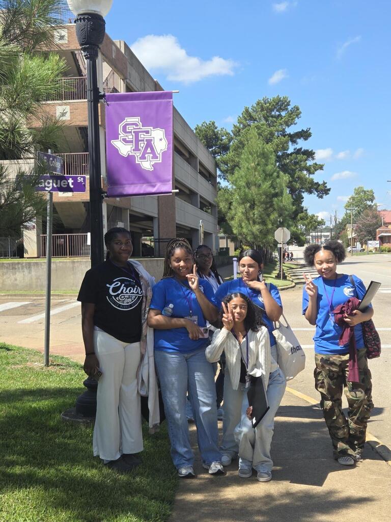 Six individuals stand near a lamp post. One holds a purple banner. A building and trees are in the background.