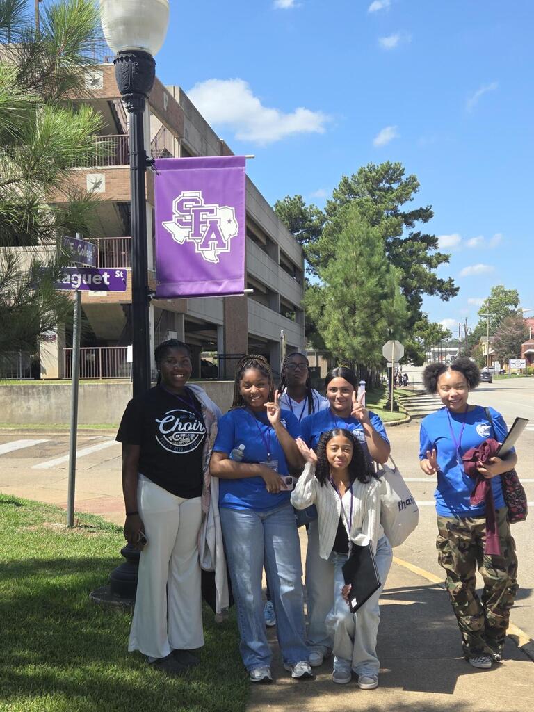 Five people in blue shirts and different colored pants posing together on a sidewalk in front of a building.
