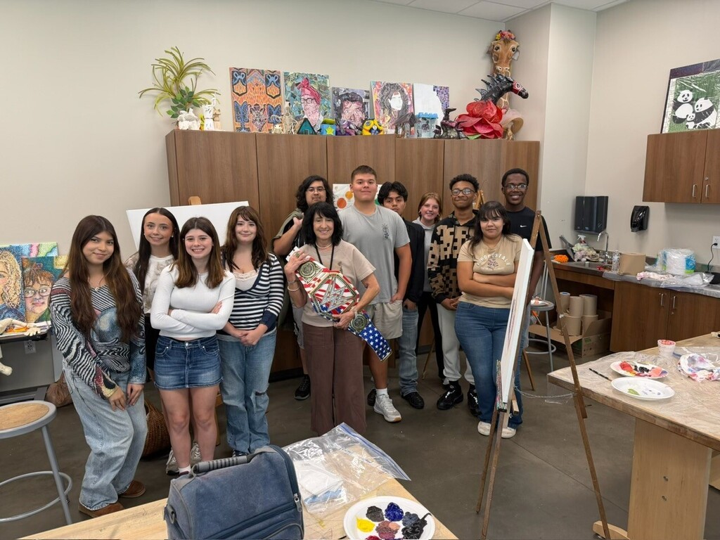 A group of people stand in a room with wooden furniture and tables. Art supplies are on the tables. Artwork is on the walls.
